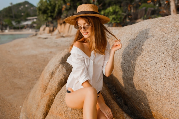 Donna bionda seducente in blusa bianca che posa sulla spiaggia tropicale