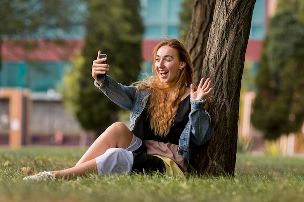Donna bionda seduta accanto a un albero e prendendo un selfie