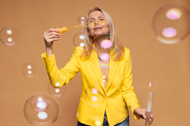 Donna bionda senior che soffia bolle su uno sfondo giallo