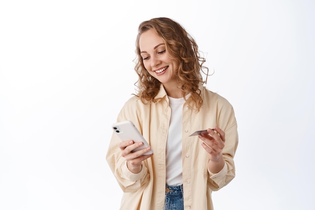 Donna bionda sorridente che paga online con carta di credito guardando il telefono cellulare in piedi su sfondo bianco Concetto di tecnologia