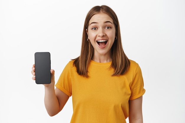 Donna bionda sorridente eccitata che mostra lo schermo, l'interfaccia dell'applicazione del telefono cellulare, la dimostrazione dell'app per smartphone, in piedi su sfondo bianco