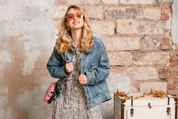 Donna bionda sorridente elegante attraente in jeans e giacca oversize che cammina contro il muro in strada