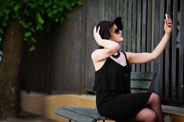 Donna bionda su collane di occhiali da sole vestito nero e cappello seduto su una panchina con il telefono cellulare a portata di mano e le cuffie