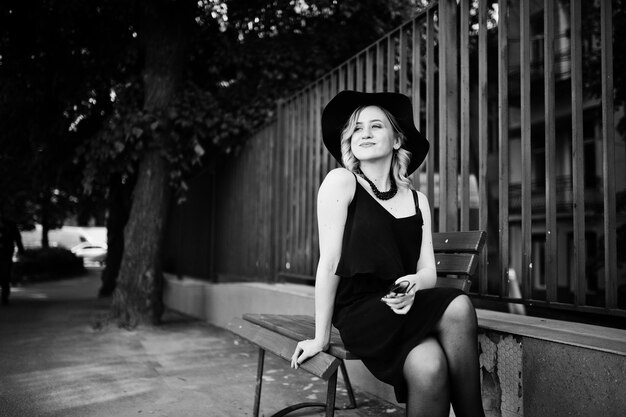 Donna bionda su collane di vestito nero e cappello seduto su una panchina