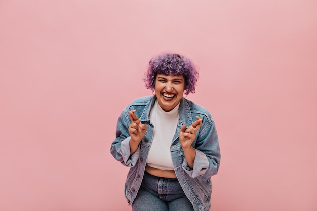 Donna brillante con capelli viola ondulati in giacca di jeans oversize ride e incrocia le dita. La donna moderna in top bianco si diverte.