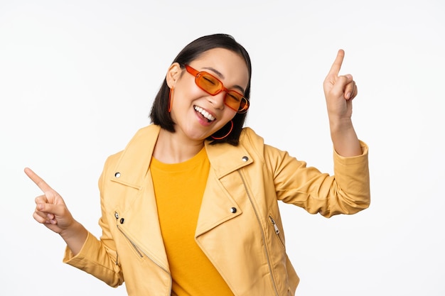 Donna bruna asiatica sorridente in occhiali da sole che punta le dita lateralmente a sinistra ea destra mostrando varianti che ridono e ballano indossando occhiali da sole sfondo bianco