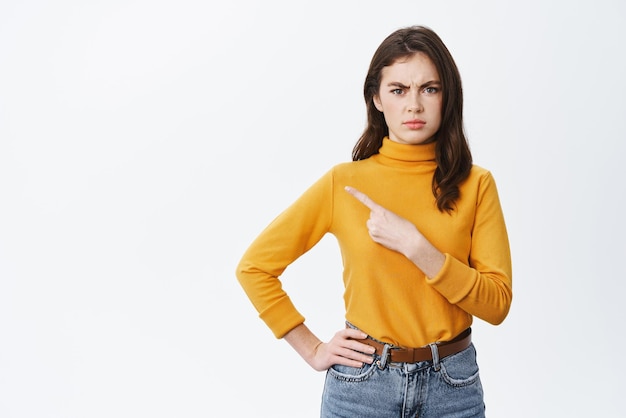 Donna bruna confusa e arrabbiata accigliata che punta il dito a sinistra con la faccia imbronciata dispiaciuta che si lamenta di qualcosa di brutto in piedi su sfondo bianco