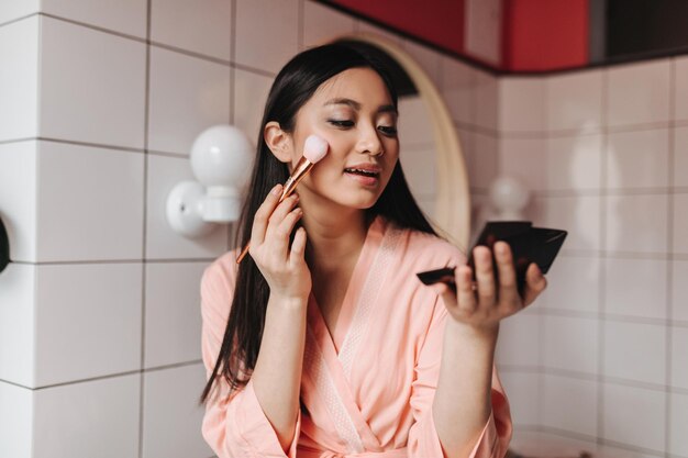 Donna bruna contorni mentre tiene piccolo specchio nero Signora in accappatoio rosa in posa in bagno