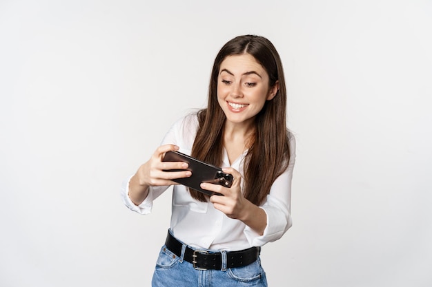 Donna bruna felice che gioca al videogioco mobile sorride e guarda lo schermo eccitata in piedi sopra w...