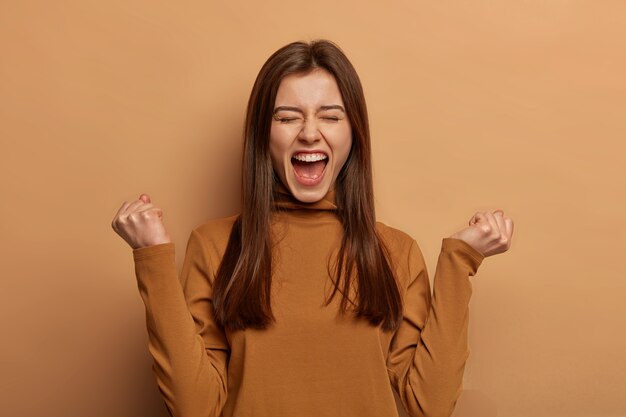 Donna bruna felicissima fa pompare i pugni, celebra il successo, grida e si sente come una campionessa, tiene la bocca spalancata, indossa un dolcevita