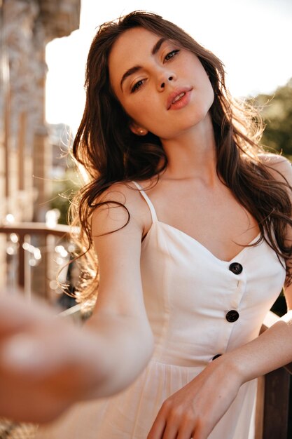 Donna bruna in abito bianco prende selfie sulla terrazza Ragazza carina con occhi marroni in abiti leggeri è fotografata sul balcone