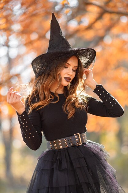 Donna bruna in costume da strega in piedi nella foresta autunnale il giorno di Halloween. Donna che indossa abiti neri e cappello a cono. Donna che posa per una foto.
