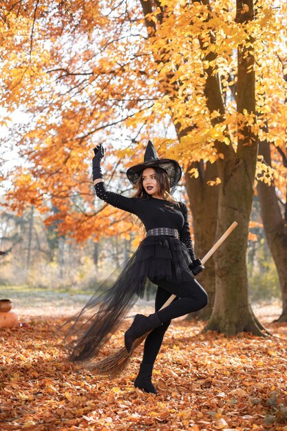 Donna bruna in costume da strega in piedi nella foresta autunnale il giorno di Halloween. Donna che indossa abiti neri e cappello a cono. Donna seduta su una scopa.