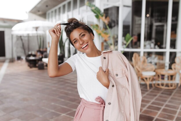 Donna bruna positiva in maglietta bianca e giacca rosa tocca la sua fascia per capelli e sorride contro il muro della terrazza del caffè