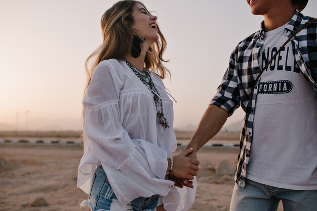 Donna bruna sognante in camicetta vintage bianca cammina con il ragazzo in camicia a scacchi e ridendo. Ritratto di bella giovane donna sorridente divertendosi in data all'aperto con un cielo incredibile