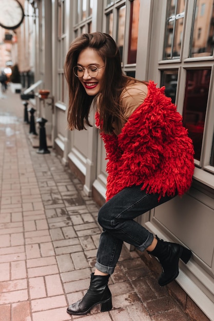 Donna carina con labbra rosse in occhiali in posa sullo sfondo della bella facciata in legno di casa con finestre Colpo a figura intera di ragazza alla moda in abito rosso