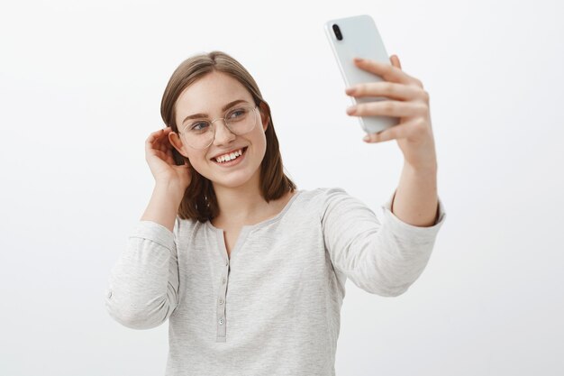 Donna carina e gentile che fa selfie da inviare all'app di appuntamenti in attesa del vero amore, sfogliando una ciocca di capelli dietro l'orecchio e sorridendo teneramente allo schermo dello smartphone in piedi femminile sopra il muro grigio