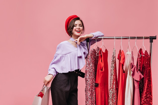 Donna carina in cappello luminoso e camicetta viola è appoggiata al supporto con abiti e posa con pacchetto su sfondo isolato.