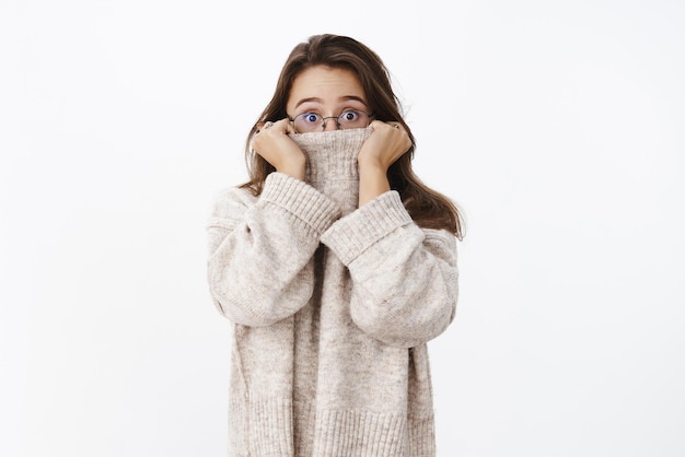 Donna carina sorpresa con gli occhiali spaventata dal freddo inverno che si nasconde nel colletto del maglione che dà una sbirciatina con gli occhi aperti alla telecamera mentre si sente scioccata e sbalordita mentre si scalda in un abito accogliente sopra il muro grigio