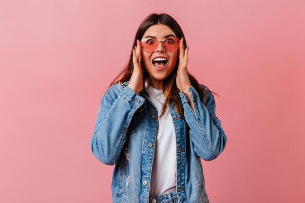 Donna carina stupita che guarda l'obbiettivo con la bocca aperta. Vista frontale della ragazza sorpresa in giacca di jeans isolato su sfondo rosa.