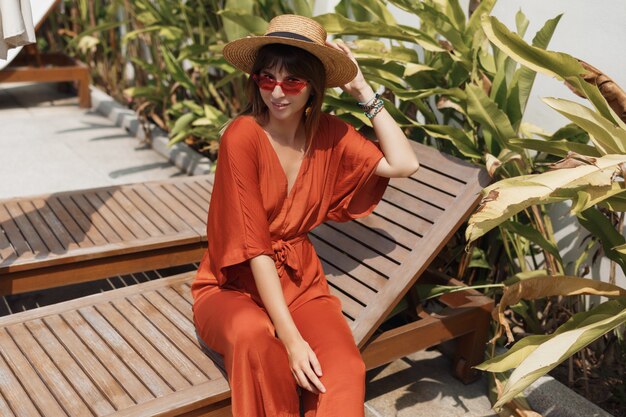 Donna castana apparente in attrezzatura arancio alla moda e cappello di paglia che si rilassano sullo sdraio vicino allo stagno.