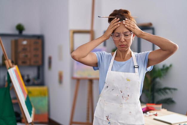Donna castana che dipinge presso uno studio d'arte che soffre di mal di testa disperata e stressata perché il dolore e l'emicrania le mani sulla testa