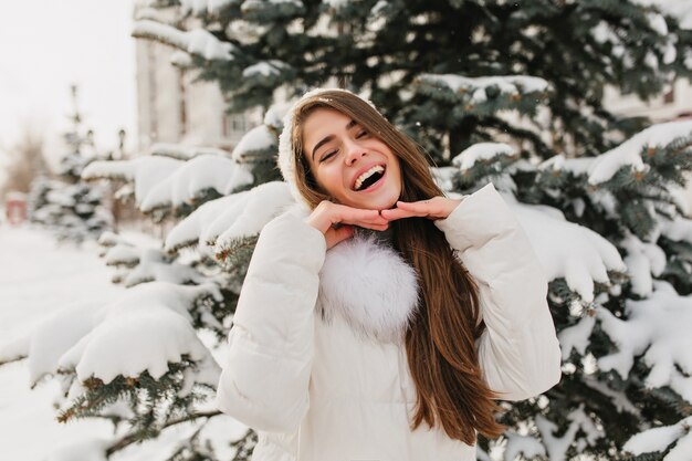 Donna castana dai capelli lunghi che posa con l'espressione del viso felice nella mattina d'inverno. Ritratto all'aperto di affascinante modello femminile europeo in cappello bianco divertendosi sull'abete innevato