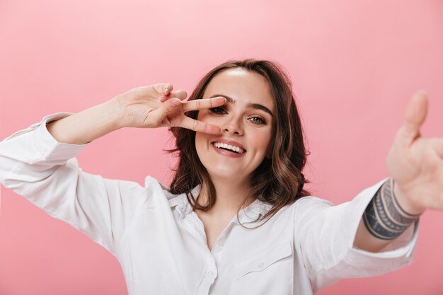 Donna castana di buon umore prende selfie su isolato. Ragazza riccia in camicia bianca sorride ampiamente e mostra il segno di pace su sfondo rosa.