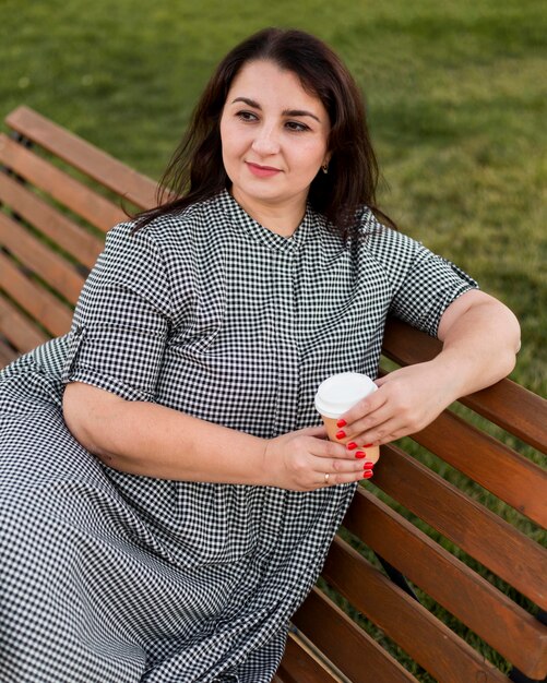 Donna castana di smiley che tiene una tazza di caffè mentre sedendosi su una panchina