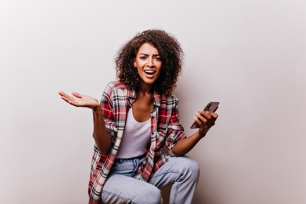 Donna castana emotiva che tiene smartphone. Meravigliosa ragazza africana in posa con il dispositivo in mano.