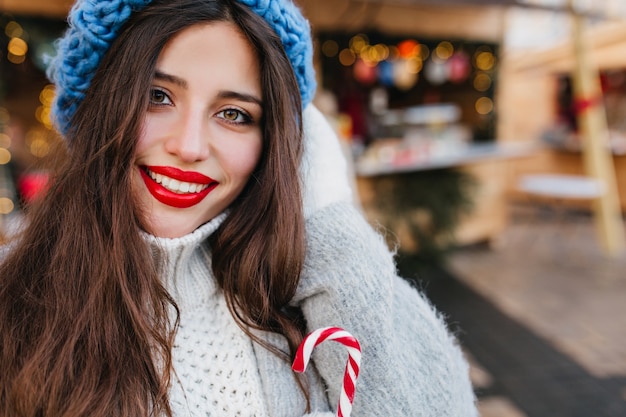 Donna castana emozionante con gli occhi marroni che bevono tè sulla strada della sfuocatura. Foto all'aperto della splendida signora dai capelli scuri in cappotto e cappello blu che tiene una tazza di caffè caldo in una giornata fredda.