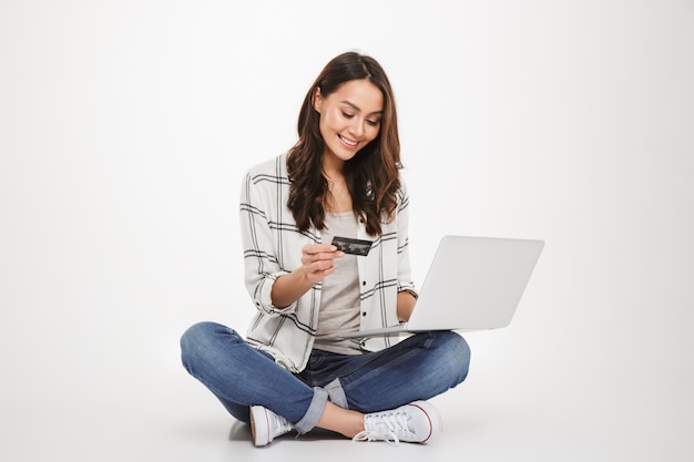 Donna castana felice verticale in camicia che si siede sul pavimento con il computer portatile e la carta di credito sopra grey