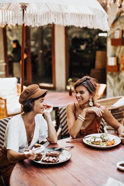 Donna castana in berretto marrone e maglietta bianca e la sua amica in elegante top sorride e riposa in street cafe