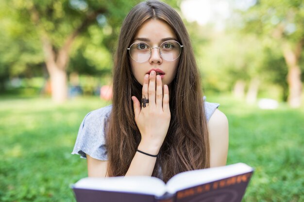 Donna castana scioccata che si siede nel parco tenendo il libro e guardando la telecamera