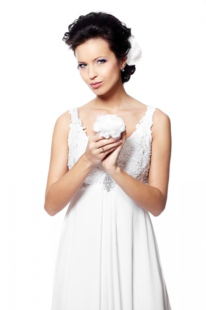 Donna castana sexy felice della bella sposa in vestito da sposa bianco con il fiore in mani con l'acconciatura e trucco luminoso con il fiore in capelli isolati su bianco