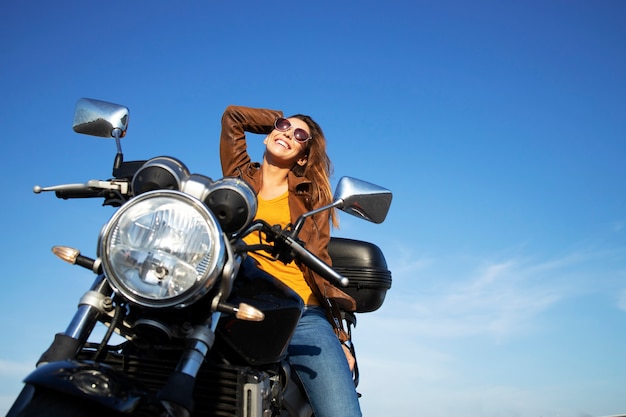 Donna castana sexy in giacca di pelle che si siede sulla moto stile retrò in una bella giornata di sole