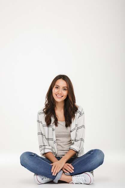 Donna castana sorridente verticale in camicia che si siede sul pavimento e che esamina la macchina fotografica sopra grey