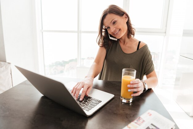 Donna casuale sorridente che parla sullo smartphone, facendo uso del computer portatile e bevendo succo dal tavolo