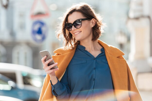Donna casuale sorridente in occhiali da sole che esaminano telefono cellulare