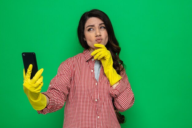Donna caucasica abbastanza premurosa delle pulizie con guanti di gomma che si mette la mano sul mento e tiene il telefono