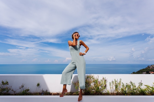 Donna caucasica abbronzata in forma in colore menta blu chiaro nel complesso sul balcone della terrazza della villa di lusso con splendida vista sul mare tropicale