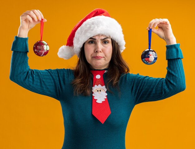 Donna caucasica adulta arrabbiata con cappello santa e cravatta santa tenendo gli ornamenti di palla di vetro isolati su sfondo arancione con spazio di copia