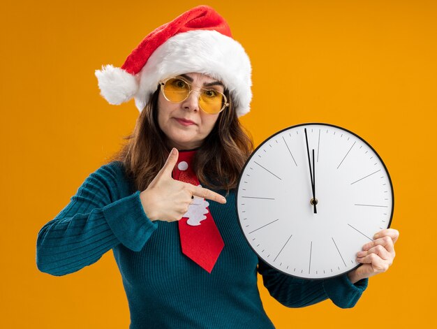 Donna caucasica adulta infastidita in occhiali da sole con cappello da Babbo Natale e cravatta di Babbo Natale che tiene e punta all'orologio isolato sulla parete arancione con spazio di copia