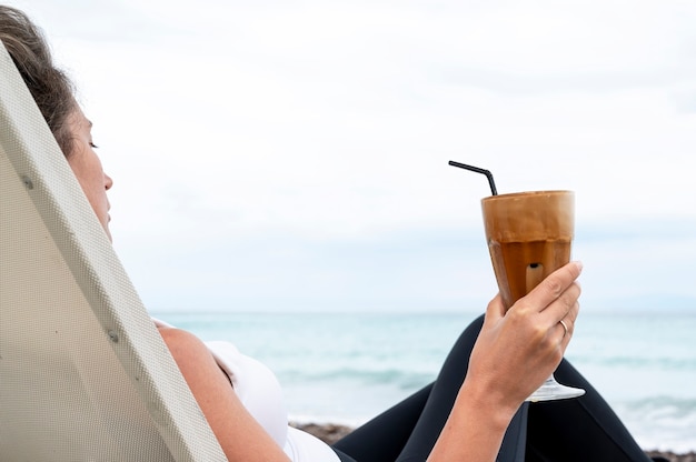 Donna caucasica che tiene la bevanda del caffè su una spiaggia con schiuma e cannuccia con il mare