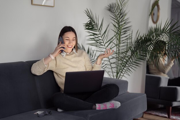 Donna caucasica che usa il laptop mentre è seduta sul divano di casa nel soggiorno serale parlando al telefono