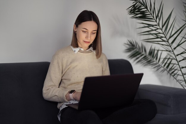 Donna caucasica che usa il laptop mentre è seduta sul divano di casa nel soggiorno serale