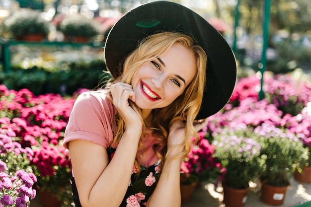 Donna caucasica estatica agghiacciante sull'aranciera al mattino. Giovane donna ispirata con cappello nero in posa con fiori rosa