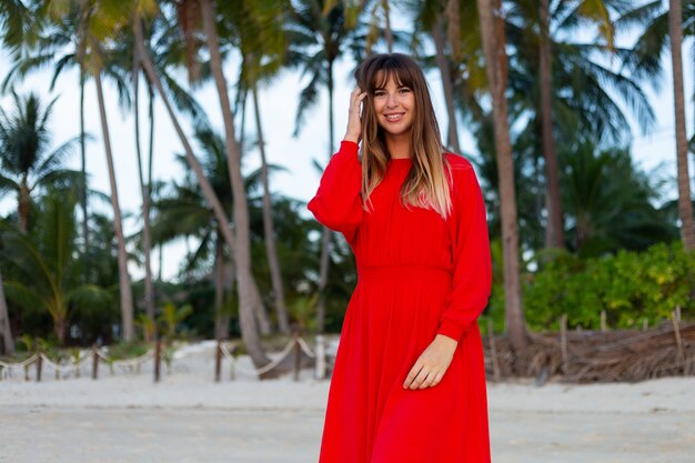 Donna caucasica in abito estivo rosso in romantico stato d'animo felice sulla spiaggia di sabbia bianca tropicale al tramonto