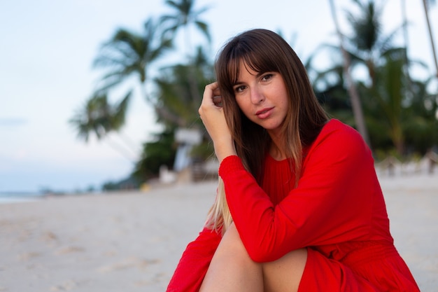 Donna caucasica in abito estivo rosso in romantico stato d'animo felice sulla spiaggia di sabbia bianca tropicale al tramonto