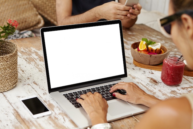 Donna caucasica in occhiali da sole, navigare in internet, visualizzare immagini e guardare video tramite i social media mentre si fa colazione al bar, seduto al tavolo di legno con computer portatile aperto